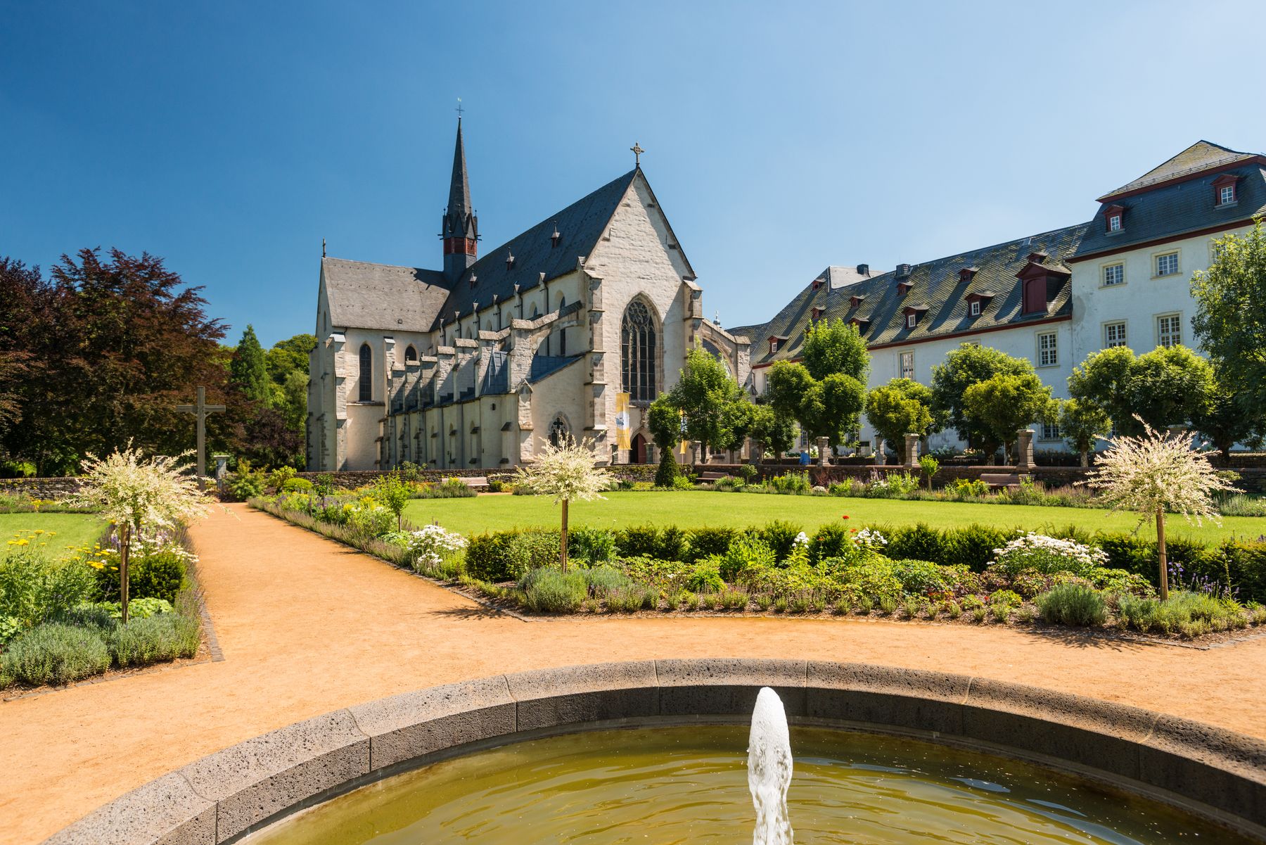 Eine historische Steinkirche mit einem hohen Kirchturm steht an einem sonnigen Tag hinter einem gepflegten Garten mit Wegen, Grünpflanzen und einem runden Brunnen im Vordergrund.